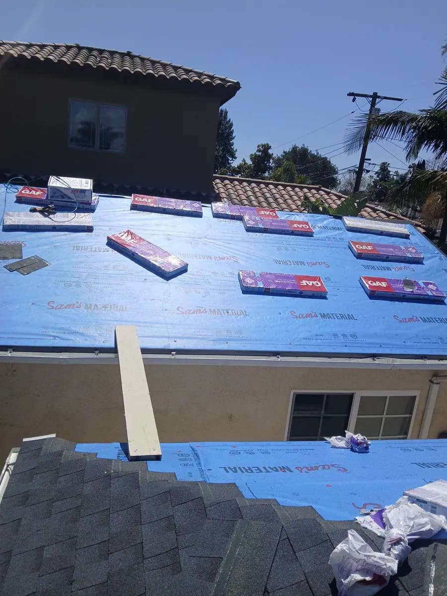 Blue roof underlayment with packaged shingles and an adjacent tile-covered roof in the background